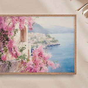 以下が含まれることがあります： ピンクの花が前景に描かれた、海岸の町の水彩画。町は青い海を見下ろす丘の中腹に建てられています。絵は明るい木製の額縁に入っています。