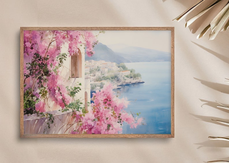 以下が含まれることがあります： ピンクの花が前景に描かれた、海岸の町の水彩画。町は青い海を見下ろす丘の中腹に建てられています。絵は明るい木製の額縁に入っています。