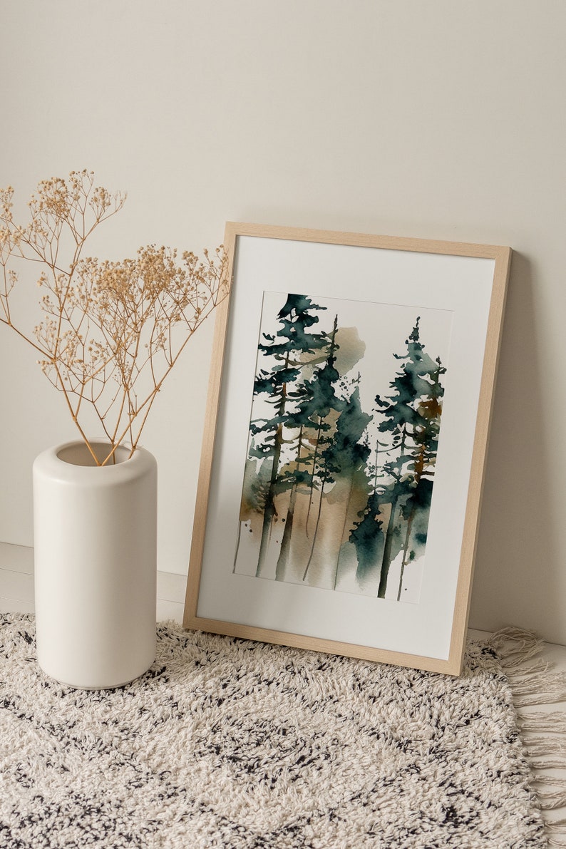 May include: Watercolor painting of a forest scene in a light wood frame. The artwork features tall, slender trees in shades of green and brown against a soft, neutral background. A white vase with dried flowers sits to the left.