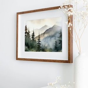 May include: A framed watercolor painting of a misty mountain landscape with a forest of pine trees in the foreground. The painting is in a brown wooden frame and is hanging on a white wall.
