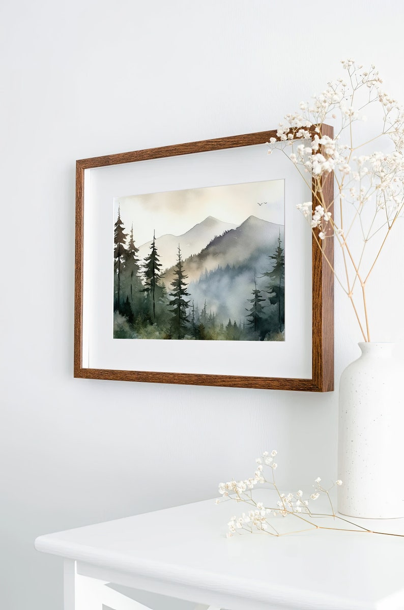 May include: A framed watercolor painting of a misty mountain landscape with a forest of pine trees in the foreground. The painting is in a brown wooden frame and is hanging on a white wall.