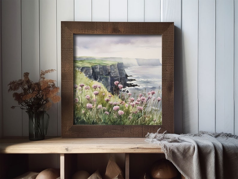 K&ouml;nnte beinhalten: Ein gerahmtes Aquarellbild einer K&uuml;stenlandschaft mit gr&uuml;nen Klippen, einem blauen Meer und rosa Wildblumen im Vordergrund.