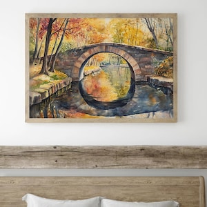 May include: Watercolor painting of a stone bridge arching over a calm river. The artwork features autumn foliage in shades of red, orange, and yellow, reflected in the water. The painting is framed in a light-colored wooden frame.