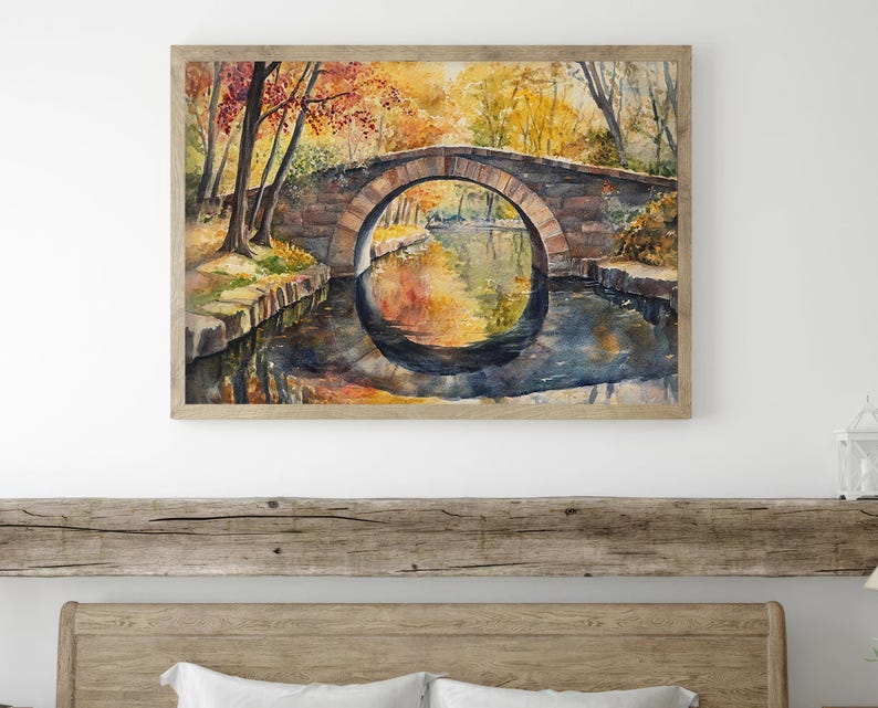 May include: Watercolor painting of a stone bridge arching over a calm river. The artwork features autumn foliage in shades of red, orange, and yellow, reflected in the water. The painting is framed in a light-colored wooden frame.