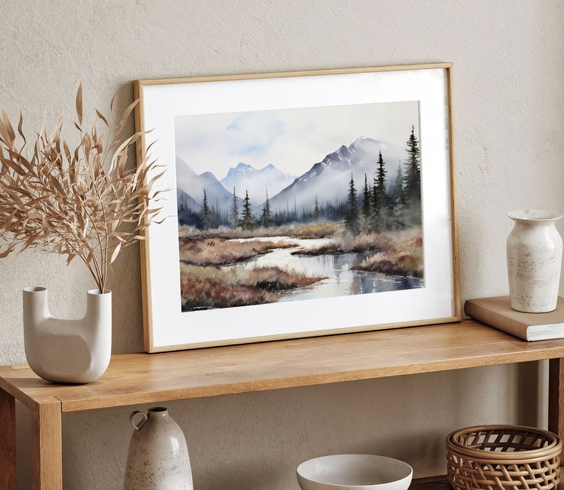 May include: A framed watercolor painting of a mountain landscape with a river flowing through it. The painting is in shades of blue, green, and brown, and features a light blue sky with white clouds.