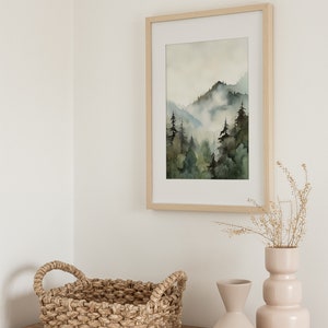 Pu&ograve; includere: Un dipinto ad acquerello incorniciato in legno che rappresenta una scena di foresta nebbiosa con alberi verdi e un cielo azzurro chiaro. Un cesto intrecciato con due manici e due vasi in ceramica rosa chiaro e beige sono su un tavolo di legno.
