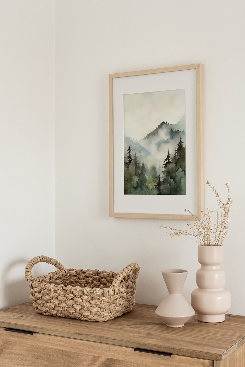 Pu&ograve; includere: Un dipinto ad acquerello incorniciato in legno che rappresenta una scena di foresta nebbiosa con alberi verdi e un cielo azzurro chiaro. Un cesto intrecciato con due manici e due vasi in ceramica rosa chiaro e beige sono su un tavolo di legno.