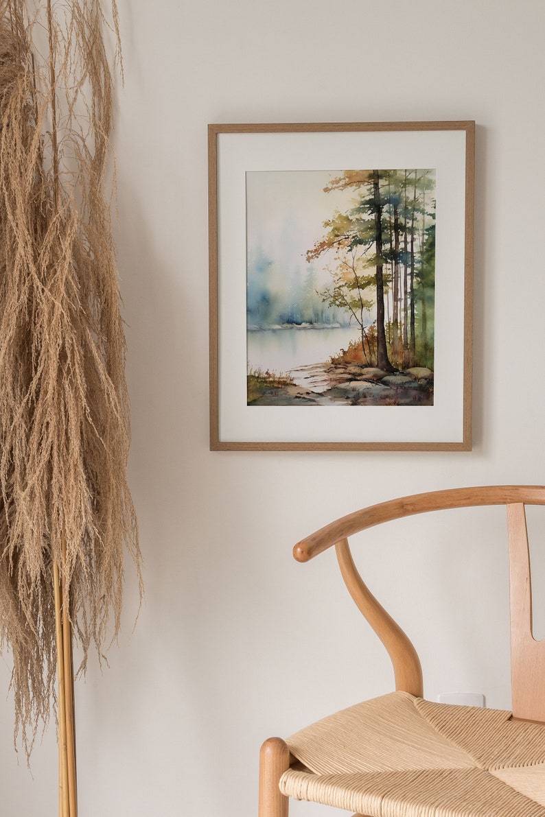 May include: A framed watercolor painting of a misty forest scene with a lake in the background. The trees are in shades of green and brown, and the water is a light blue. The painting is set against a white wall.