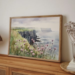 K&ouml;nnte beinhalten: Ein Aquarellbild einer Klippe, die &uuml;ber ein Gew&auml;sser blickt. Die Klippen sind dunkelgrau und das Wasser ist hellblau. Im Vordergrund befinden sich rosa Blumen.