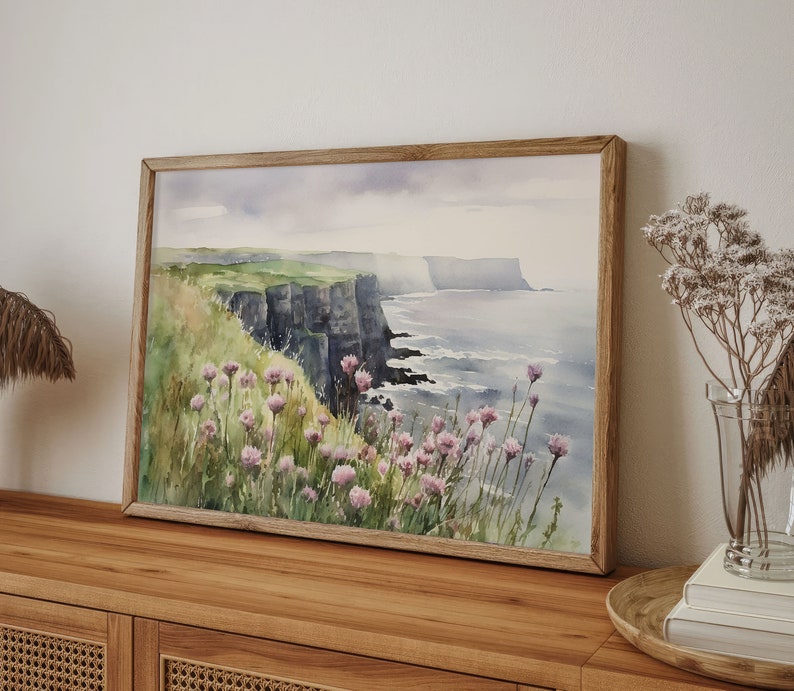 K&ouml;nnte beinhalten: Ein Aquarellbild einer Klippe, die &uuml;ber ein Gew&auml;sser blickt. Die Klippen sind dunkelgrau und das Wasser ist hellblau. Im Vordergrund befinden sich rosa Blumen.