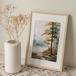 May include: A framed watercolor painting of a forest scene with a lake in the background. The painting is in shades of green, brown, and blue. The frame is made of light wood.