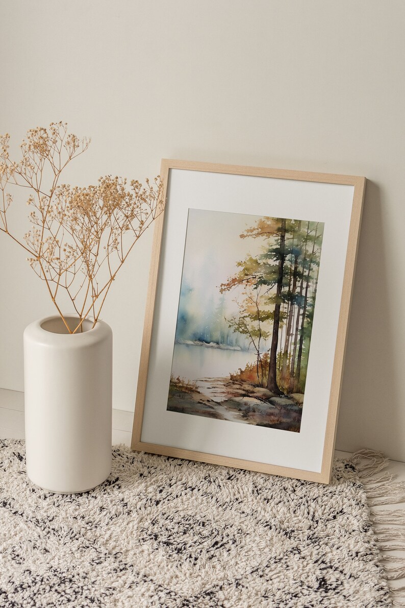 May include: A framed watercolor painting of a forest scene with a lake in the background. The painting is in shades of green, brown, and blue. The frame is made of light wood.