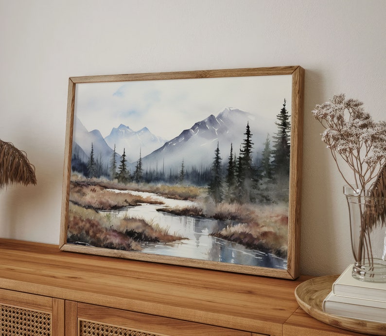 May include: A framed watercolor painting depicting a mountain landscape with a stream flowing through the foreground. The mountains are painted in shades of blue and gray, and the trees are a dark green. The painting is set against a white background.