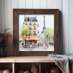 May include: Watercolor print of a Parisian street scene in a brown wooden frame. The artwork depicts a cafe with an orange awning, a building, the Eiffel Tower, and trees. The print is displayed on a wooden shelf with a vase and a blanket.