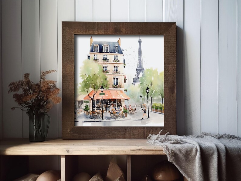 May include: Watercolor print of a Parisian street scene in a brown wooden frame. The artwork depicts a cafe with an orange awning, a building, the Eiffel Tower, and trees. The print is displayed on a wooden shelf with a vase and a blanket.