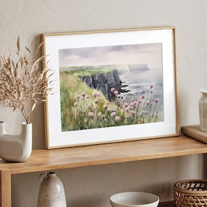 K&ouml;nnte beinhalten: Ein gerahmtes Aquarellbild einer Klippe, die &uuml;ber ein Gew&auml;sser blickt. Das Bild zeigt eine gr&uuml;ne Grasklippe mit rosa Wildblumen im Vordergrund. Die Klippe ist dunkelgrau und erstreckt sich in die Ferne. Das Wasser ist hellblau und der Himmel ist hellgrau.