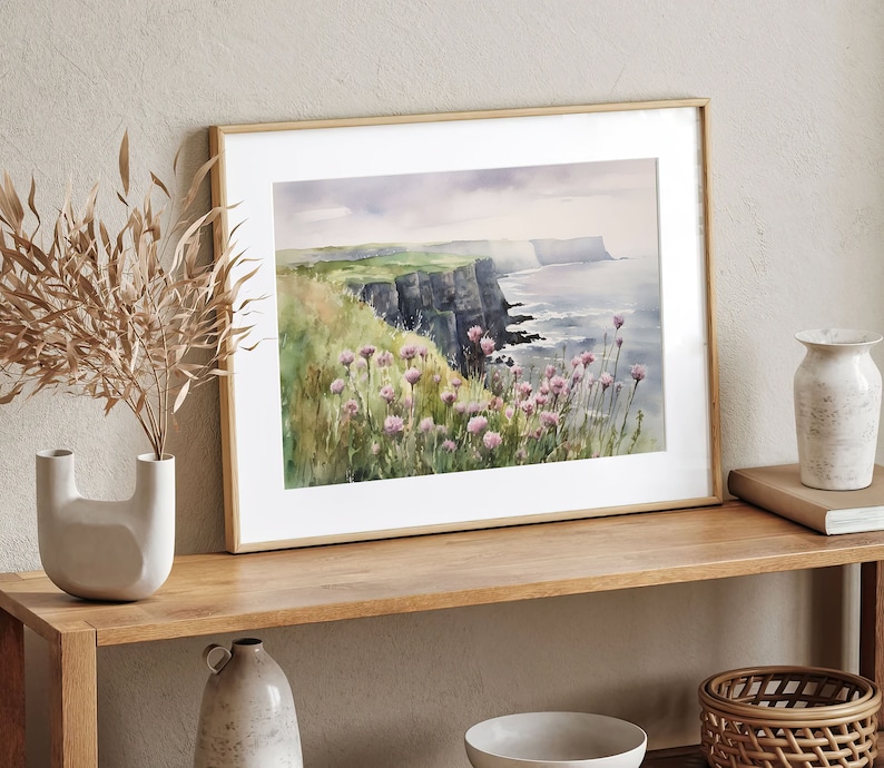 K&ouml;nnte beinhalten: Ein gerahmtes Aquarellbild einer Klippe, die &uuml;ber ein Gew&auml;sser blickt. Das Bild zeigt eine gr&uuml;ne Grasklippe mit rosa Wildblumen im Vordergrund. Die Klippe ist dunkelgrau und erstreckt sich in die Ferne. Das Wasser ist hellblau und der Himmel ist hellgrau.