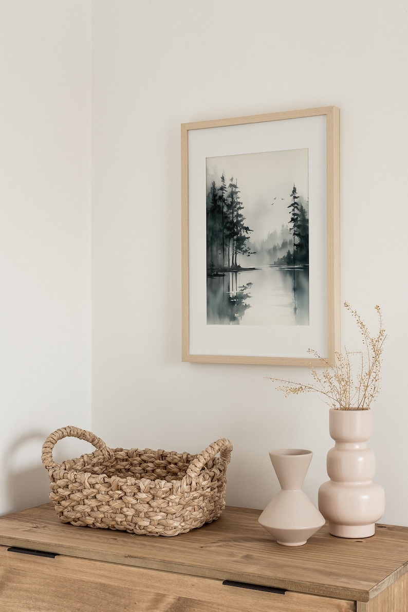 May include: A framed watercolor painting of a lake scene with trees and a misty background. The painting is hanging on a white wall above a wooden dresser with a woven basket and two ceramic vases.