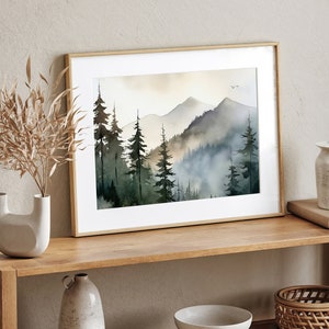 May include: A framed watercolor painting of a misty mountain landscape with a forest of dark green trees. The painting is in a light wood frame and is hanging on a wall.