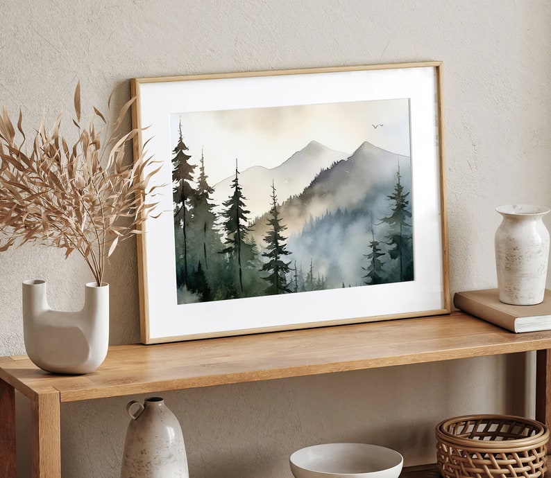 May include: A framed watercolor painting of a misty mountain landscape with a forest of dark green trees. The painting is in a light wood frame and is hanging on a wall.