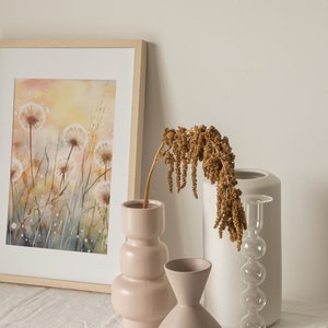 May include: A framed watercolor painting of a field of dandelions with a soft, yellow and blue background. The painting is displayed on a table with three vases, one with dried flowers, one with a pink textured design, and one with a white textured design.