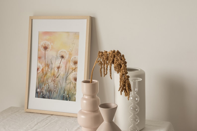 May include: A framed watercolor painting of a field of dandelions with a soft, yellow and blue background. The painting is displayed on a table with three vases, one with dried flowers, one with a pink textured design, and one with a white textured design.