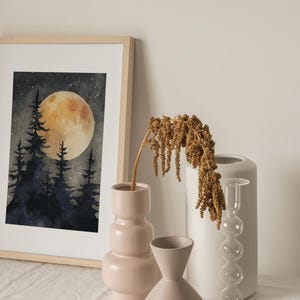 May include: A framed watercolor painting depicts a nighttime forest scene with a large, glowing yellow moon. Silhouetted trees are in the foreground. Beside the painting are decorative vases in shades of pink and white, and a dried plant.