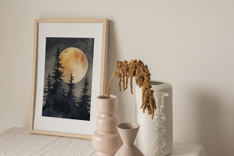May include: A framed watercolor painting depicts a nighttime forest scene with a large, glowing yellow moon. Silhouetted trees are in the foreground. Beside the painting are decorative vases in shades of pink and white, and a dried plant.