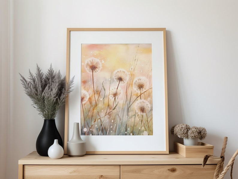 May include: A framed watercolor painting of a field of white dandelions with a soft yellow and orange background. The painting is in a light wood frame and is hanging on a white wall.