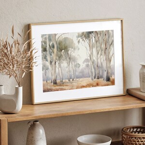 May include: A framed watercolour painting depicting a forest scene with tall, slender trees in shades of grey and brown. The trees are set against a pale blue sky with a hint of green foliage in the background. The painting is displayed on a wooden shelf with other decorative items.