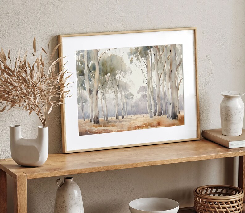 May include: A framed watercolour painting depicting a forest scene with tall, slender trees in shades of grey and brown. The trees are set against a pale blue sky with a hint of green foliage in the background. The painting is displayed on a wooden shelf with other decorative items.
