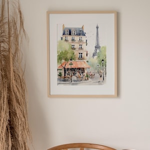 May include: Watercolor painting of a Parisian street scene with a cafe, buildings, and the Eiffel Tower. The artwork is framed in a light wood frame and hangs on a neutral wall. The painting features soft colors and captures the essence of Paris.