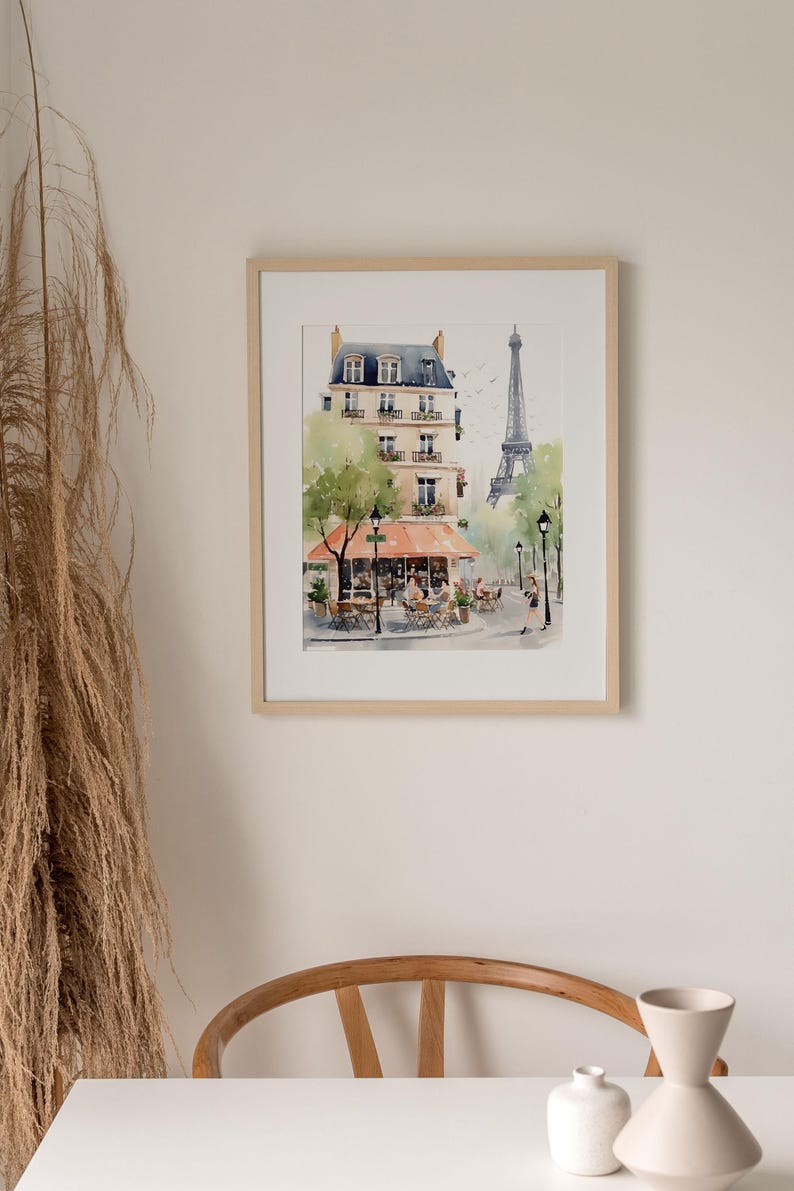 May include: Watercolor painting of a Parisian street scene with a cafe, buildings, and the Eiffel Tower. The artwork is framed in a light wood frame and hangs on a neutral wall. The painting features soft colors and captures the essence of Paris.