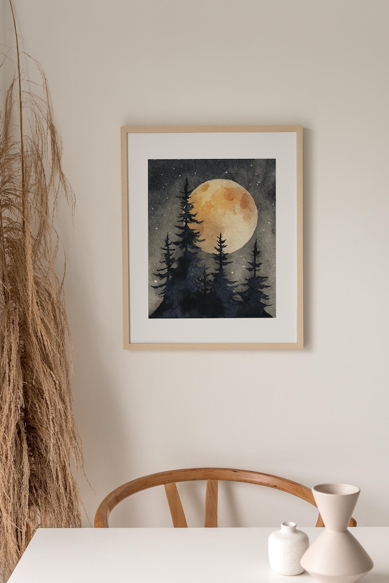 May include: Watercolor painting of a night scene with a large, golden moon and silhouetted pine trees. The artwork is framed in a light wood frame and hangs on a neutral-colored wall. The overall theme is nature and landscape.