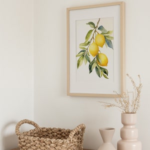 Op de afbeelding: Een ingelijst aquarel schilderij van een citroen tak met drie citroenen die eraan hangen. Het schilderij is op een witte achtergrond en is ingelijst in een licht houten lijst. Het schilderij staat op een houten tafel met een geweven mand en twee keramische vazen.