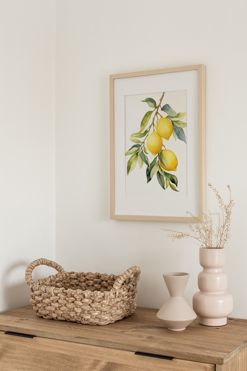 Op de afbeelding: Een ingelijst aquarel schilderij van een citroen tak met drie citroenen die eraan hangen. Het schilderij is op een witte achtergrond en is ingelijst in een licht houten lijst. Het schilderij staat op een houten tafel met een geweven mand en twee keramische vazen.