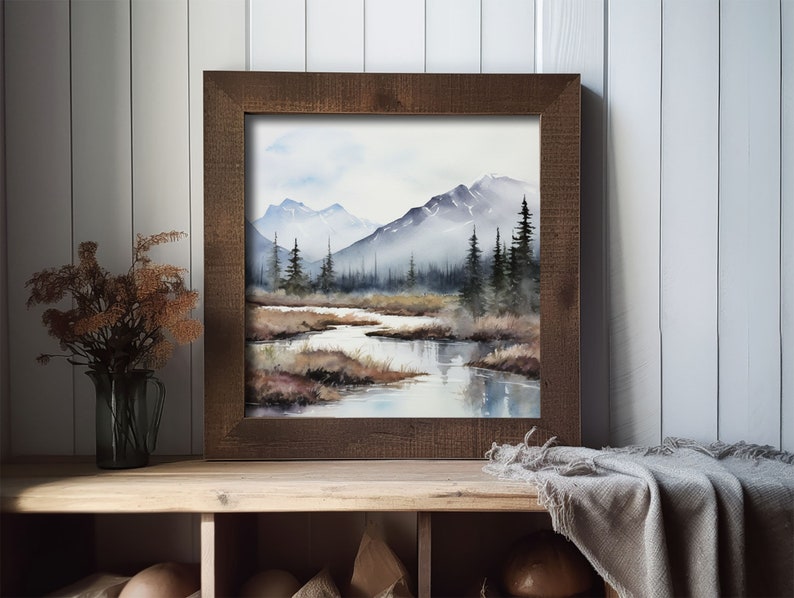 May include: A framed watercolor painting of a mountain landscape with a river flowing through the foreground. The mountains are in the background and are covered in a light mist. The river is a light blue color and reflects the sky. The trees are a dark green color and are scattered throughout the landscape.