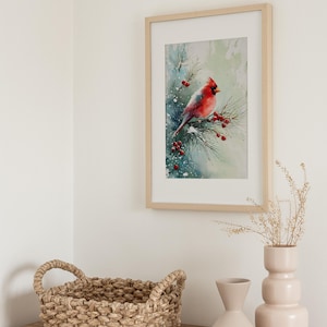 Op de afbeelding: Een ingelijste aquarelprint van een rode kardinaal op een dennentak met rode bessen. Het kunstwerk staat in een licht houten lijst, boven een houten kast met een gevlochten mand en twee lichtroze vazen.