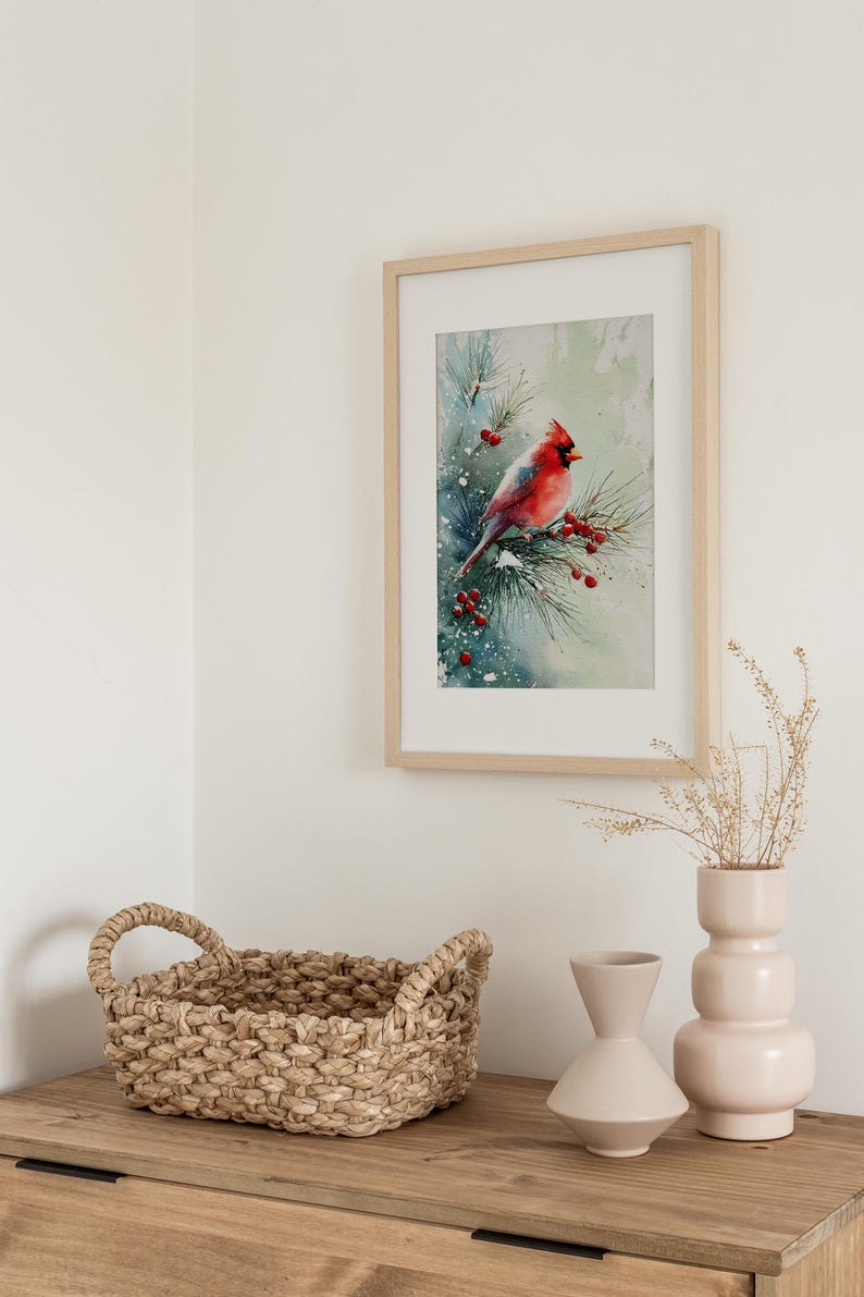 Op de afbeelding: Een ingelijste aquarelprint van een rode kardinaal op een dennentak met rode bessen. Het kunstwerk staat in een licht houten lijst, boven een houten kast met een gevlochten mand en twee lichtroze vazen.