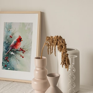 Op de afbeelding: Een ingelijste aquarelprint van een rode kardinaal die op een besneeuwde tak met rode bessen zit. Het kunstwerk staat in een licht houten lijst en wordt tentoongesteld met decoratieve vazen.