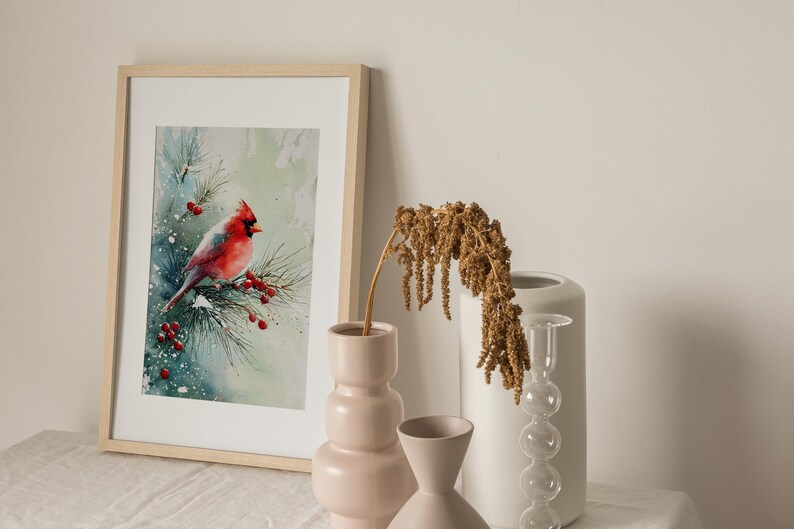 Op de afbeelding: Een ingelijste aquarelprint van een rode kardinaal die op een besneeuwde tak met rode bessen zit. Het kunstwerk staat in een licht houten lijst en wordt tentoongesteld met decoratieve vazen.