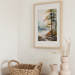 May include: A framed watercolor painting of a lake scene with trees in the background. The painting is hanging on a white wall above a wooden table with a woven basket and two ceramic vases.