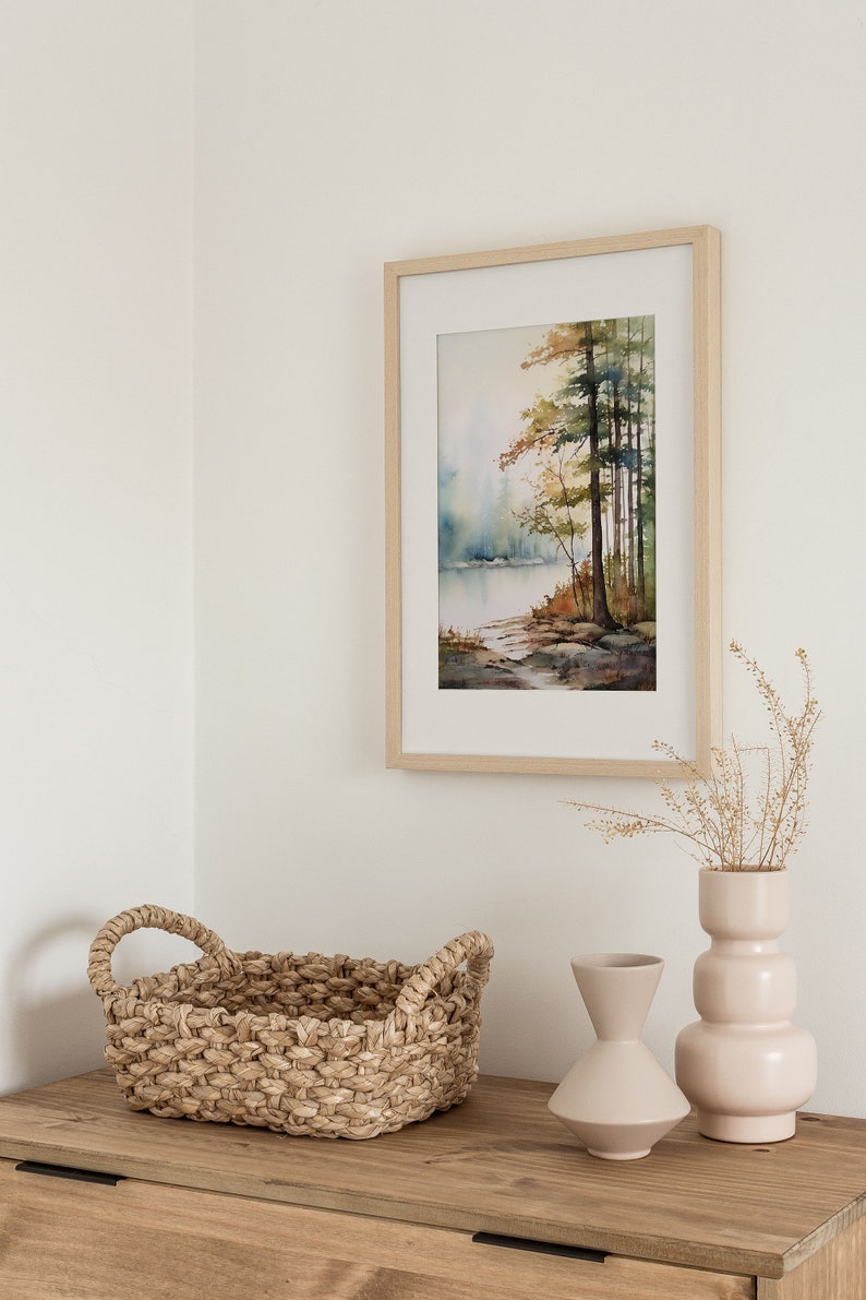 May include: A framed watercolor painting of a lake scene with trees in the background. The painting is hanging on a white wall above a wooden table with a woven basket and two ceramic vases.