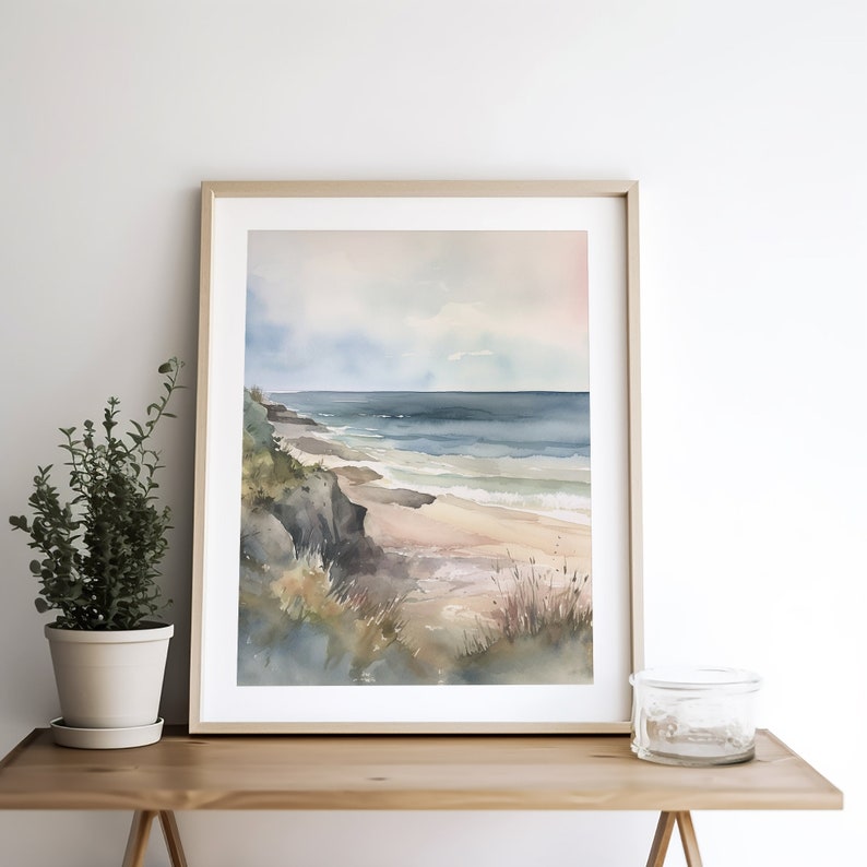 May include: A watercolor painting of a seascape with a sandy beach, blue ocean, and a rocky cliff. The painting is framed in a light wood frame and is hanging on a white wall.