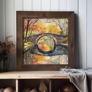 May include: Watercolor painting of a stone bridge over a calm river, framed in a dark wood. The artwork depicts autumn foliage with warm colors reflected in the water. The painting is displayed on a wooden shelf.