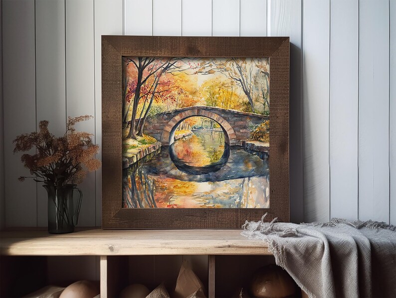 May include: Watercolor painting of a stone bridge over a calm river, framed in a dark wood. The artwork depicts autumn foliage with warm colors reflected in the water. The painting is displayed on a wooden shelf.