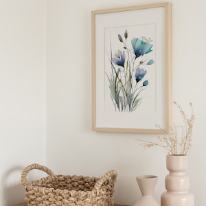May include: A framed watercolor painting of blue flowers with green stems and leaves. The painting is on a white wall above a wooden table with a woven basket, a beige vase with dried flowers, and a light beige vase.