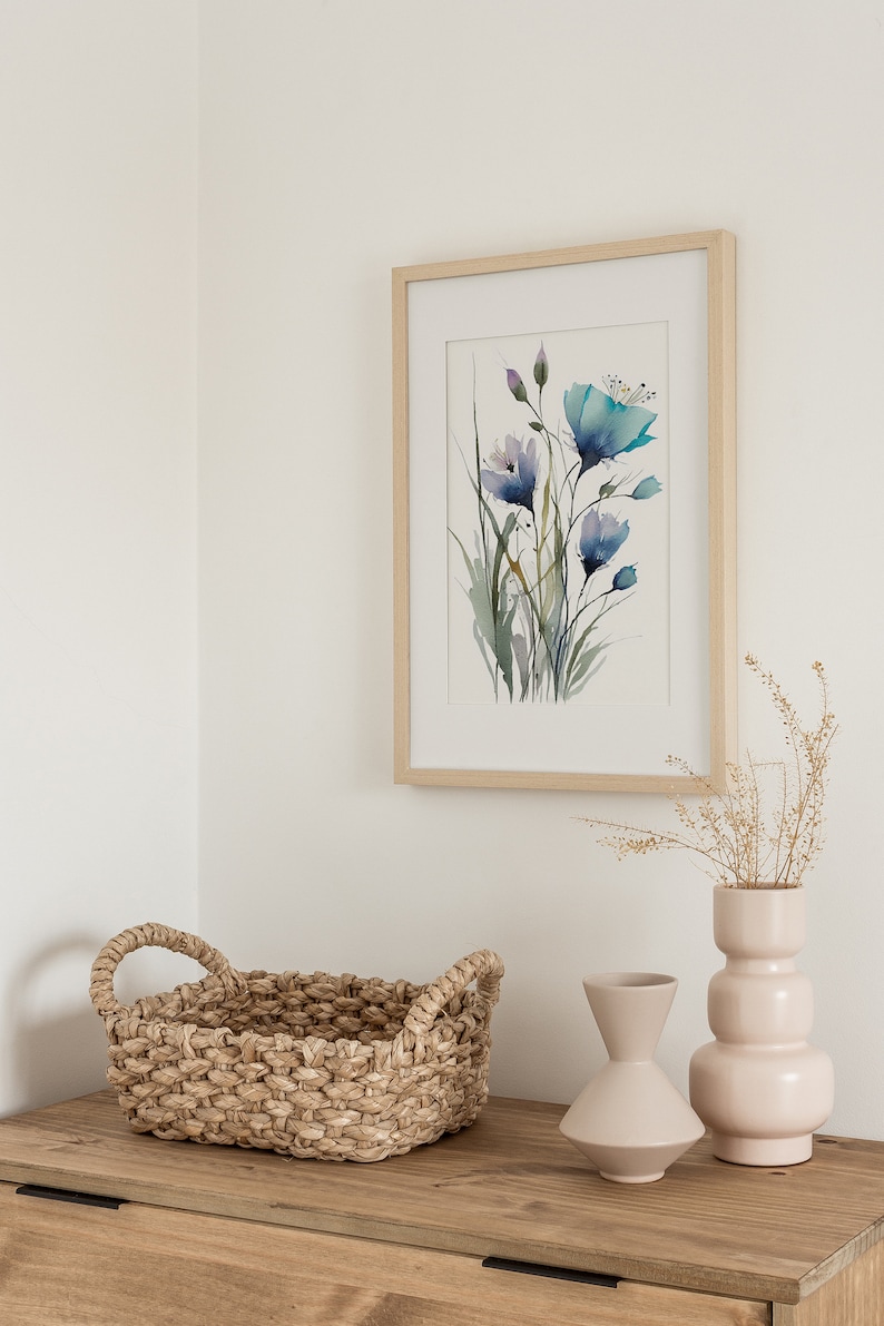 May include: A framed watercolor painting of blue flowers with green stems and leaves. The painting is on a white wall above a wooden table with a woven basket, a beige vase with dried flowers, and a light beige vase.