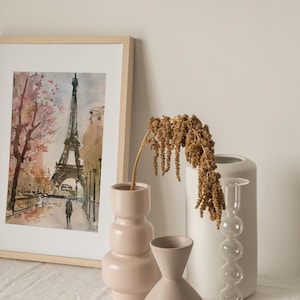 Puede incluir: Una pintura de acuarela enmarcada de la Torre Eiffel en Par&iacute;s, Francia. La pintura representa la torre con un &aacute;rbol rosa y blanco en primer plano. La pintura se exhibe sobre una superficie blanca con tres jarrones.