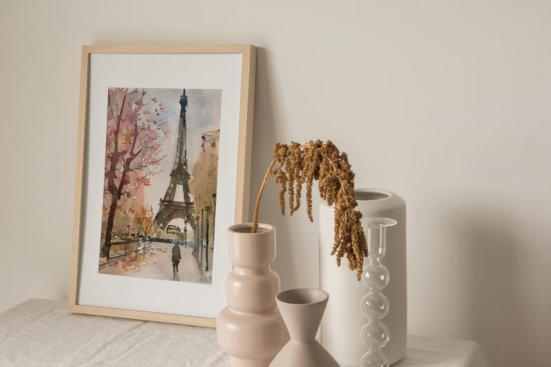 Puede incluir: Una pintura de acuarela enmarcada de la Torre Eiffel en Par&iacute;s, Francia. La pintura representa la torre con un &aacute;rbol rosa y blanco en primer plano. La pintura se exhibe sobre una superficie blanca con tres jarrones.